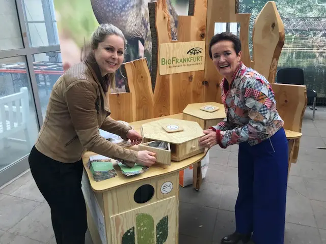 Zwei Frauen stehen an einem Holztisch mit einem drehbaren Fach und einem Schild mit der Aufschrift BioFrankfurt.