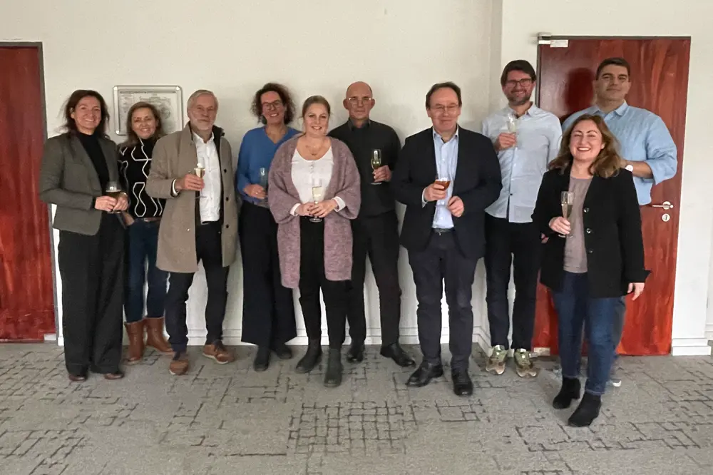 Gruppenfoto von elf Personen in festlicher Atmosphäre, stehend mit Sektgläsern vor heller Wand und zwei geschlossenen Türen – Anlass: eine Kooperationsvereinbarung.