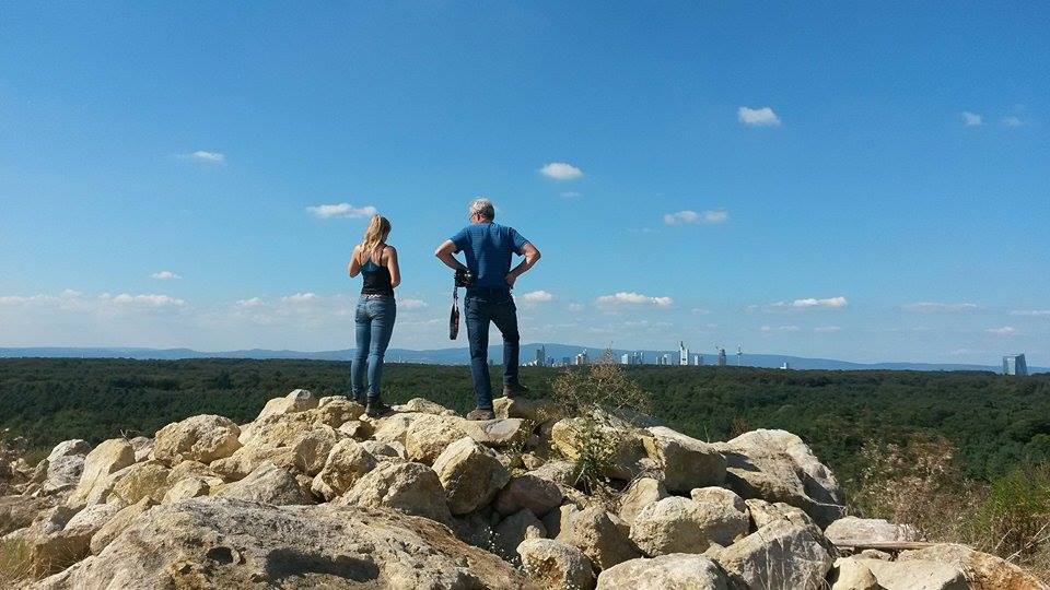 Zwei Personen stehen auf einem Felsenhaufen und blicken auf einen bewaldeten Hügel mit einer Stadt im Hintergrund unter blauem Himmel.