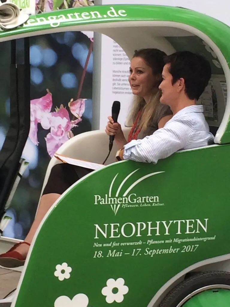 Zwei Frauen sitzen in einem grünen Fahrzeug mit der Aufschrift PalmenGarten Neophyten und einem Veranstaltungsdatum von Mai bis September 2017.