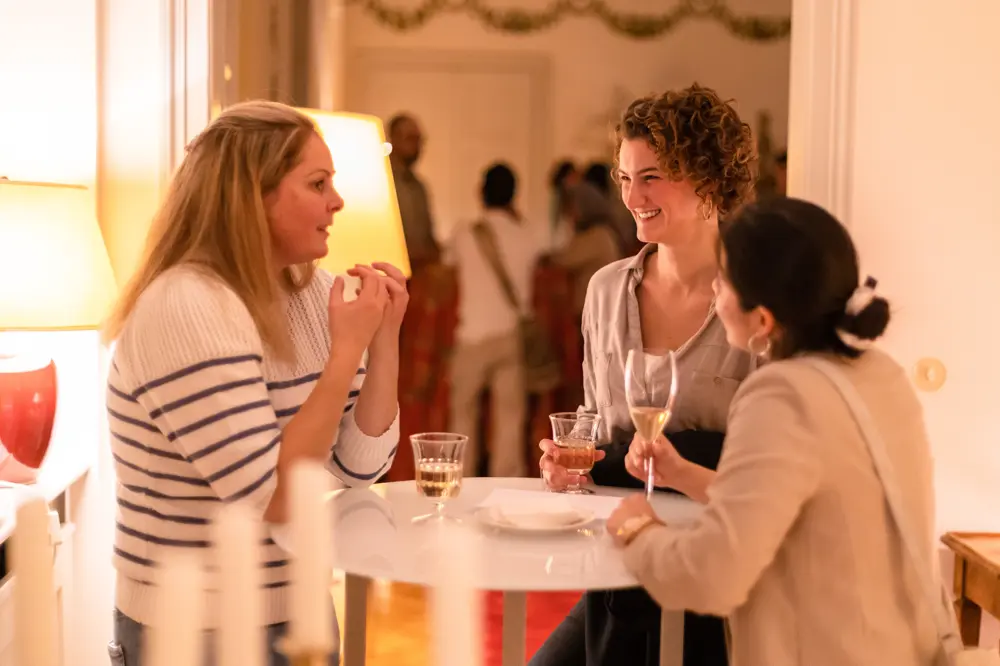Drei Frauen im Gespräch an einem Stehtisch bei einem Empfang oder einer Abendveranstaltung, mit Weingläsern in der Hand, im Hintergrund weitere Gäste in warm beleuchtetem Raum.