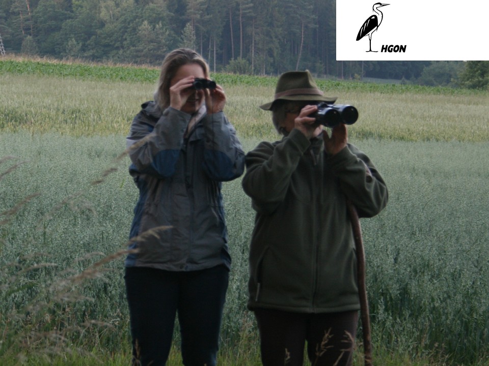 Eine junge Frau mit Fernglas steht neben einer älteren Frau mit Fernglas in der Dämmerung auf einem Feld